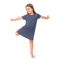 A young girl with light brown hair is posing in a slate blue short-sleeve t-shirt dress, balancing on one leg with arms outstretched against a clean white background.