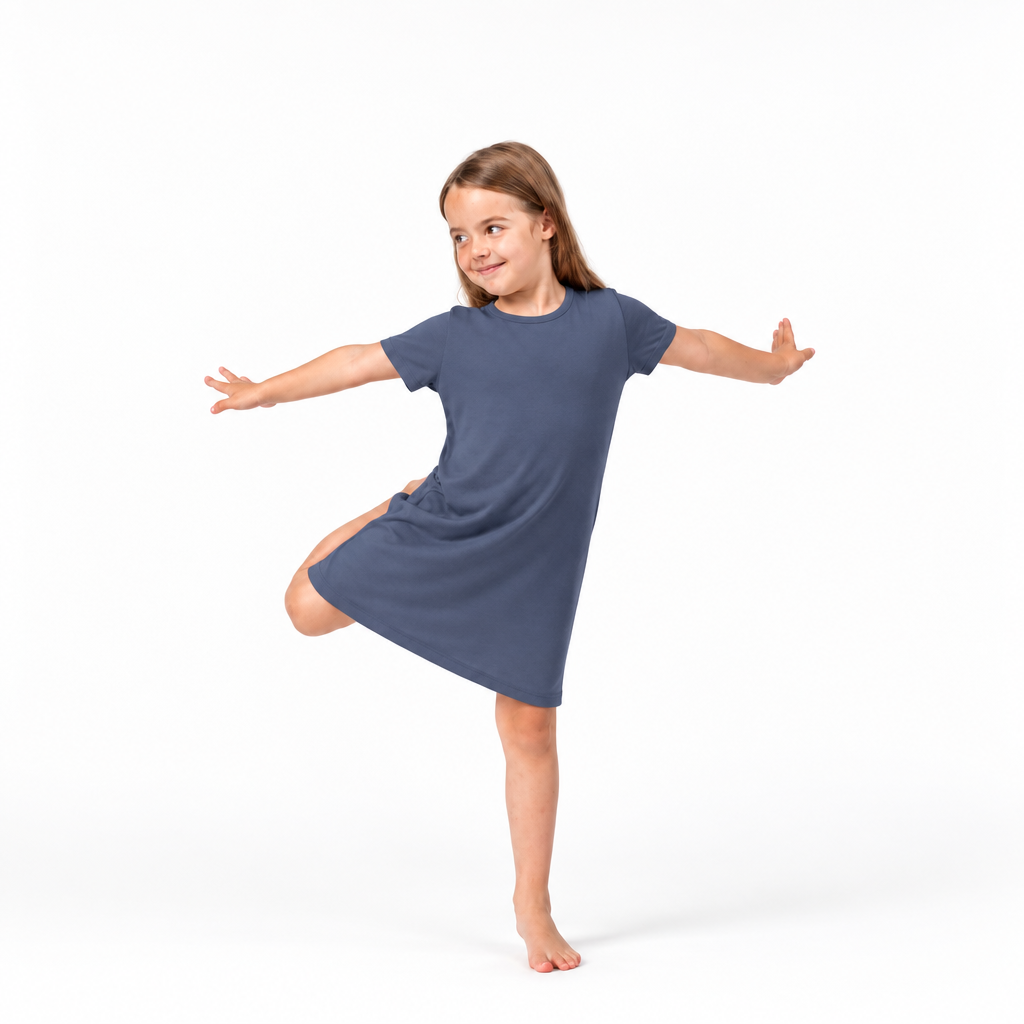 A young girl with light brown hair is posing in a slate blue short-sleeve t-shirt dress, balancing on one leg with arms outstretched against a clean white background.